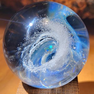 Pode incluir: Uma esfera de vidro transparente com um design azul e branco em espiral, assemelhando-se a uma onda. A esfera assenta numa pequena base de madeira quadrada. O interior apresenta detalhes iridescentes.