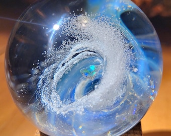 May include: A clear glass sphere with a swirling blue and white design, reminiscent of a wave. The sphere is mounted on a small, square wooden base. The interior has iridescent details.
