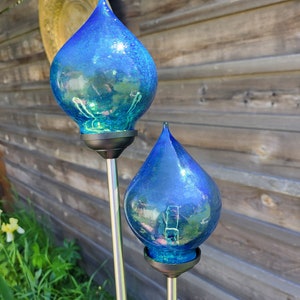May include: Two blue glass teardrop-shaped garden stakes with metal poles. The glass is iridescent and reflects light.