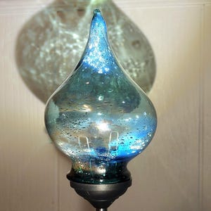 May include: A decorative glass garden stake with a teardrop-shaped blue and teal glass top. The glass has a textured, bubbly appearance and sits atop a black base and metal pole. The shadow of the glass is visible in the background.