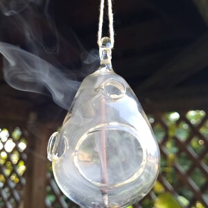 May include: A clear glass incense burner with a round opening hangs from a white string. Smoke rises from the incense stick inside the burner.