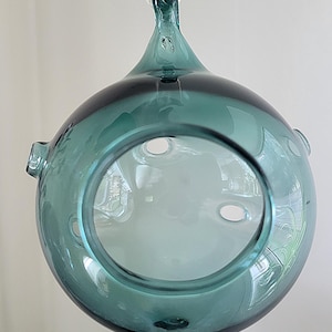 May include: A teal glass orb with a hole in the center and a loop at the top for hanging. The orb has a frosted glass interior and is perfect for air plants.