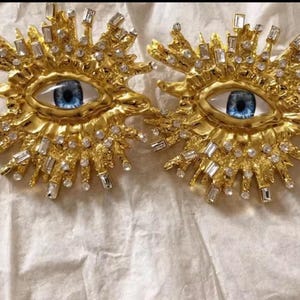 May include: A pair of gold-tone earrings with a large blue eye design. The eyes are surrounded by a halo of small, clear rhinestones and larger, baguette-shaped rhinestones.