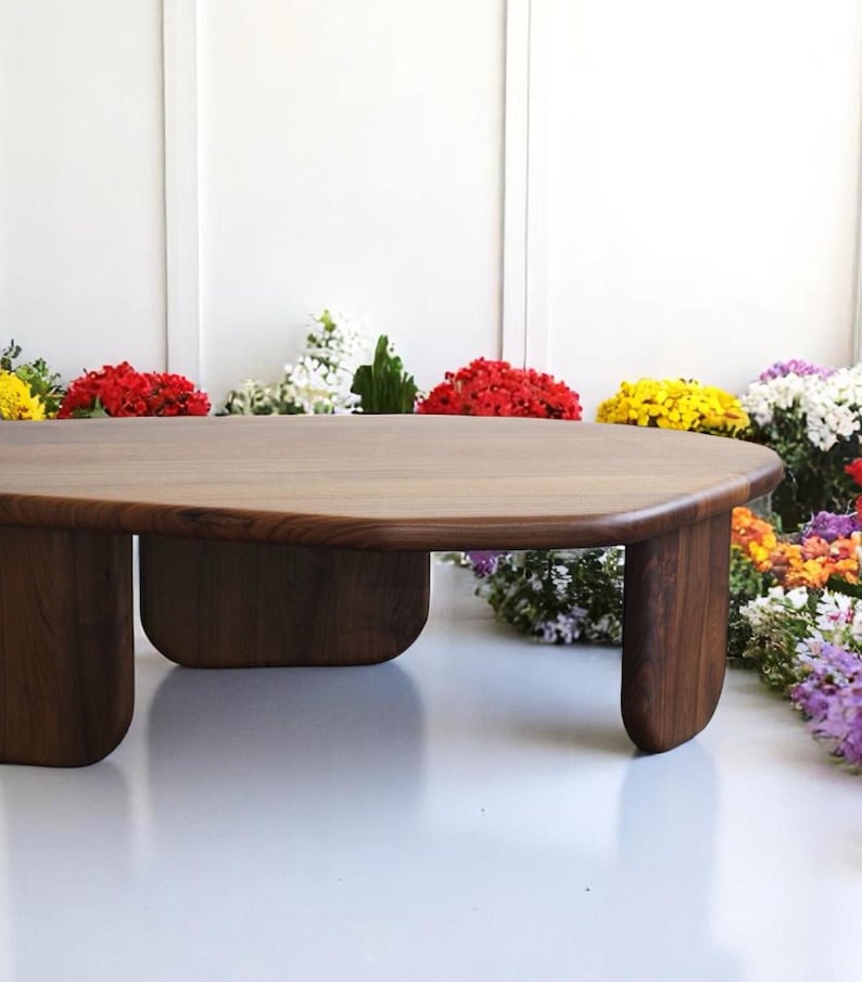 Op de afbeelding: Een donkerbruine houten salontafel met een unieke ovale vorm en vier poten. De tafel is omringd door kleurrijke bloemen.