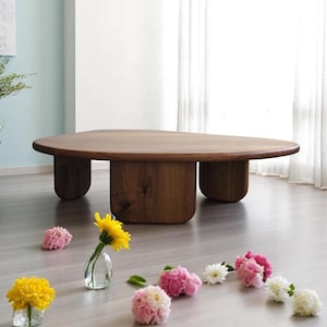 Op de afbeelding: Een grote, ovale houten salontafel met vier unieke, afgeronde poten. De tafel is gemaakt van donker hout en heeft een natuurlijke, rustieke afwerking. De tafel is omringd door een strooiing van roze en gele bloemen in kleine glazen vazen.