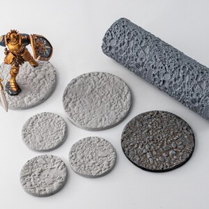 Op de afbeelding: Een grijze cilindrische textuurroller met een rotsachtig patroon, vijf ronde basissen met getextureerde oppervlakken en een geschilderd miniatuurfiguur in gouden harnas met een speer en schild. De roller heeft de tekst LUM op de zijkant.