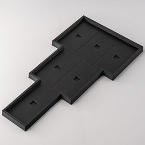 Op de afbeelding: Een zwart sieraden display dienblad met meerdere rechthoekige compartimenten. De lade heeft een trapsgewijs ontwerp, met verschillende niveaus en maten van compartimenten. Elk compartiment heeft een kleine vierkante uitsparing.