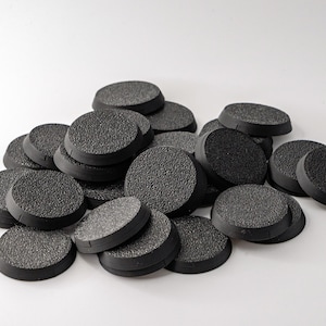 Può includere: Un mucchio di basi nere, rotonde e strutturate per miniature. Le basi hanno un bordo rialzato e una superficie ruvida, adatte per il modellismo e i giochi da tavolo. Le basi sono realizzate in un materiale grigio scuro.