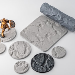 Può includere: Una collezione di basi in miniatura grigie e testurizzate in varie forme circolari e quadrate, un mattarello cilindrico grigio con un motivo roccioso e una miniatura di cavaliere dipinta in oro e blu con lancia e scudo.