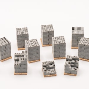 Op de afbeelding: Miniatuur stapels grijze betonblokken op houten pallets zijn gerangschikt op een wit oppervlak. Een klein figuur van een persoon in een blauw overhemd en broek staat in de buurt. De scène suggereert constructie of bouw.