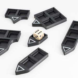 Peut inclure: Cinq composants de plateau de jeu en plastique noir avec un nombre variable de compartiments carrés et une icône de triangle blanc sur l'extrémité pointue, disposés autour d'un dé à six faces crème avec des points noirs dans l'une des pièces.