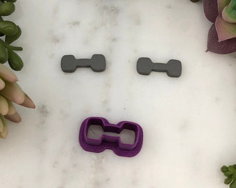 May include: Three gray plastic dumbbell cookie cutters and one purple plastic dumbbell cookie cutter on a white marble surface.
