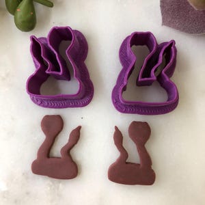 May include: Two purple plastic cookie cutters in a unique shape, along with two brown cookies in the same shape. The cutters have a textured border. The cookies are a dark brown color.