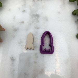 May include: A purple plastic rocket cookie cutter and a white clay rocket.