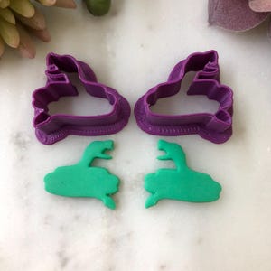 May include: Two purple plastic cookie cutters in the shape of a dinosaur head and two green dinosaur-shaped cookies. The cutters have a textured border. The cookies are a bright green color.