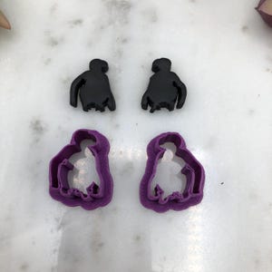 May include: Two black, silhouette-style cookie cutters and two purple cookie cutters in the shape of a person. The cutters are arranged on a white marble surface. The black cutters are solid, while the purple cutters have a detailed outline.