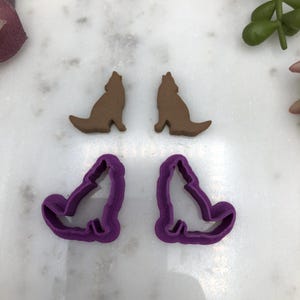 May include: Two brown wolf cookie cutters and two purple wolf cookie cutters. The cookie cutters are on a white marble surface.