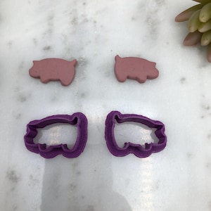 May include: Two pink pig cookie cutters and two purple pig cookie cutters. The cookie cutters are on a white marble surface.