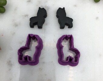 May include: Four llama-shaped cookie cutters. Two are solid black, and two are purple with a detailed outline. The cutters are made of plastic and are designed for baking or crafting. The items are arranged on a white marble surface.
