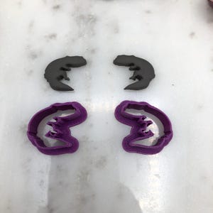 以下が含まれることがあります： 4つのパーツ：2つの濃い灰色のサンショウウオ型のオブジェクトと、2つの紫色のサンショウウオ型のクッキーカッター。オブジェクトは白い大理石の表面に正方形に配置されています。クッキーカッターは波状のデザインです。