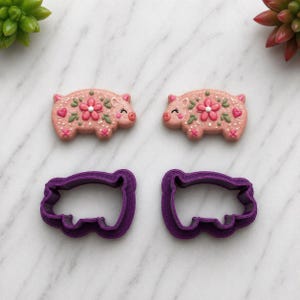 May include: Two decorated pink pig-shaped cookies with floral designs and two purple pig-shaped cookie cutters. The cookies have white sprinkles, pink flowers, green leaves, and red hearts. The cookie cutters are made of plastic.