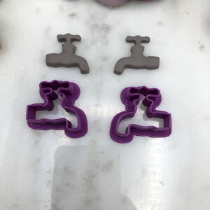 May include: Four tap-shaped items. Two are gray, solid, and three-dimensional. The other two are purple, plastic, and have a tap-shaped outline. The items are arranged on a white, marble-like surface. The items are likely cookie cutters.