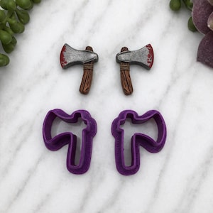 May include: Two miniature axes with silver blades and brown handles, one with red accents, are displayed above two purple axe-shaped cookie cutters. The items are arranged on a white marble surface.