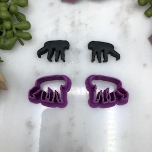 May include: Four pieces: two black bear-shaped objects and two purple bear-shaped cookie cutters. The cookie cutters have a bear shape with a ruffled edge. The objects are on a white surface with green and pink accents.