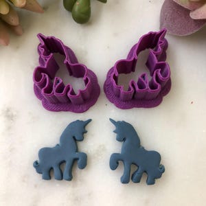 May include: Two purple plastic unicorn-shaped cookie cutters and two blue unicorn-shaped fondant decorations. The cookie cutters have a ruffled edge. The unicorns are standing with their heads up and horns pointed forward. The background is a white marble surface.