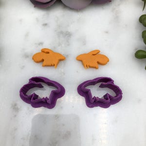 May include: Two orange bunny-shaped cookie cutters and two purple bunny-shaped cookie cutters are displayed on a white marble surface. The orange bunnies are cut-out cookies. The purple cutters are plastic.