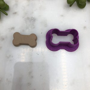 May include: A tan dog bone-shaped cookie cutter next to a purple cookie cutter. The cookie cutter is made of plastic and is designed to cut out dog bone shapes from dough or fondant. The tan dog bone is next to the purple cutter.