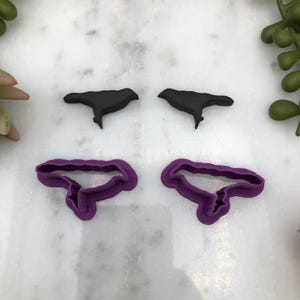 May include: Two black crow cookie cutters and two purple crow cookie cutters. The cookie cutters are on a white marble surface.
