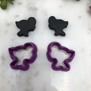 May include: Four cookie cutters in the shape of a bird. Two are solid black, and two are purple outlines. The cutters are arranged on a white marble surface.