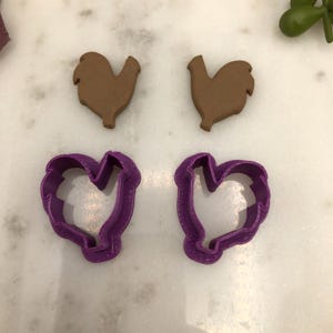 May include: Two brown rooster-shaped cookies and two purple heart-shaped cookie cutters are displayed on a white surface. The cookie cutters have a textured edge. The cookies are a solid brown color.