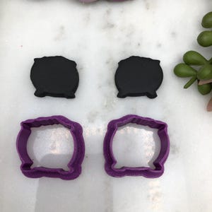 Könnte beinhalten: Zwei schwarze, kesselartige Polymer-Tonstücke und zwei lila, kesselförmige Ausstecher. Die Ausstecher haben einen strukturierten, wellenförmigen Rand. Die Artikel sind auf einer weißen Oberfläche mit einer grünen Pflanze im Hintergrund angeordnet.