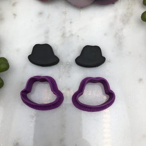 Könnte beinhalten: Zwei violette Plastik-Ausstecher in Hutform und zwei schwarze Fondanthüte.