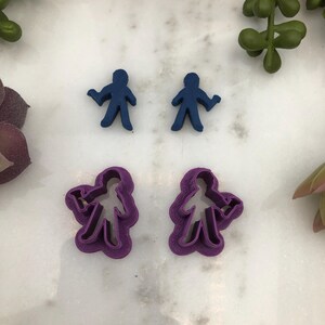May include: Two sets of cookie cutters, one set is blue and the other is purple. Both sets are shaped like stick figures holding a small object in their right hand.