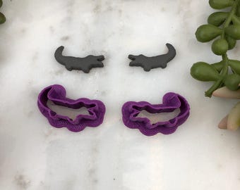May include: Two sets of alligator cookie cutters. One set is gray and the other is purple.
