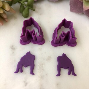 May include: Four purple polymer clay cutters in the shape of a sumo wrestler. Two cutters are the outline of the wrestler, and two are the solid shape. The cutters are on a white marble surface.