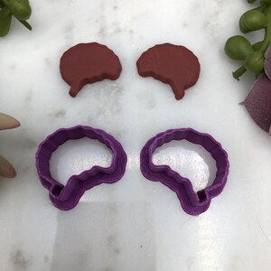 May include: Two purple plastic brain-shaped cookie cutters and two brown clay brain shapes.