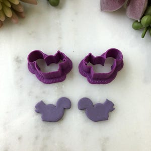 May include: Two purple squirrel-shaped cookie cutters and two squirrel-shaped fondant cutouts. The cookie cutters have a detailed border. The cutouts are a solid color and have a simple design.
