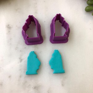 Op de afbeelding: Twee paarse plastic koekjesvormpjes in de vorm van een gestileerd silhouet van een persoon, vergezeld door twee bijpassende turquoise klei-uitsparingen die de vorm van de vormpjes weerspiegelen, met een duidelijke omtrek en een volle vorm.