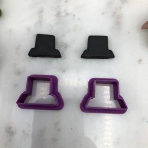 May include: Two black fondant toppers and two purple plastic cookie cutters shaped like hats. The toppers are slightly larger than the cutters.