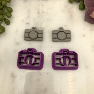 May include: Four camera-shaped cookie cutters. Two are grey and solid, while the other two are purple with an open design. The cutters are arranged on a white marble surface, suggesting a baking or crafting theme.