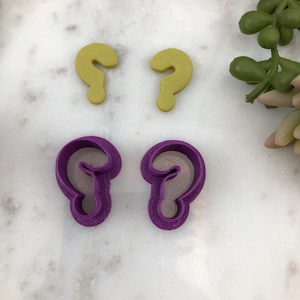 Könnte beinhalten: Zwei violette Plastik-Ausstecher in Form eines Fragezeichens und zwei gelbe Ausstecher in Form eines Fragezeichens auf einer weißen Marmorfläche.