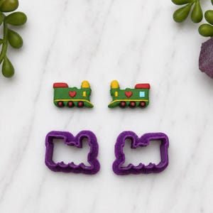 May include: Two colourful train-shaped cookie cutters and two decorated biscuits. The biscuits are green, red, and yellow with heart and circle details. The cutters are purple and have a scalloped edge.