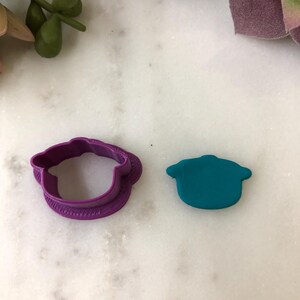 Könnte beinhalten: Ein lila Polymer Clay Ausstecher und eine türkisfarbene Polymer Clay Form. Der Ausstecher hat die Form einer Kochmütze mit dekorativem Rand. Die türkisfarbene Form ist eine Kochmütze.