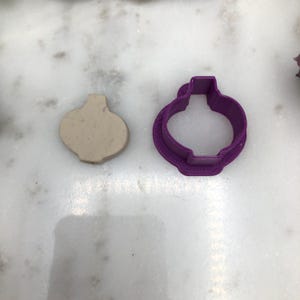 Può includere: Un tagliapasta di plastica viola a forma di ornamento, accanto a un pezzo di pasta da modellare beige.