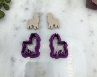 May include: Two purple llama cookie cutters and two beige llama cookie cutters. The cookie cutters are on a white marble surface.