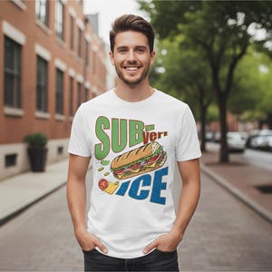 May include: White t-shirt featuring a graphic of a submarine sandwich and the words "SUBverICE" in green and blue. The design includes a detailed illustration of the sandwich with various toppings. The person is standing on a brick-paved street.